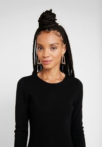 Maglione nero a maniche lunghe, texture a coste, scollo rotondo. Acconciatura intrecciata con un chignon, orecchini geometrici dorati. Sfondo neutro.