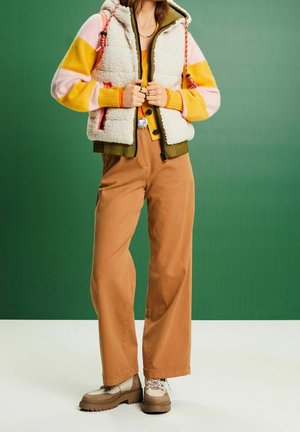 Osoba v béžové fleecové vestě přes žlutý, růžový a oranžový svetr, hnědé široké kalhoty a béžové šněrovací boty na zeleném pozadí.