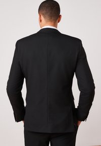 Giacca da completo nera, vestibilità sartoriale, spacco singolo sulla schiena, tessuto liscio, maniche lunghe con bottoni metallici ai polsini, design pulito.