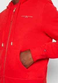 Sweat à capuche rouge avec fermeture éclair, en tissu doux, comprenant une poche avant, une fermeture éclair métallique et un logo brodé sur la poitrine. Poignets avec détails d'accentuation.