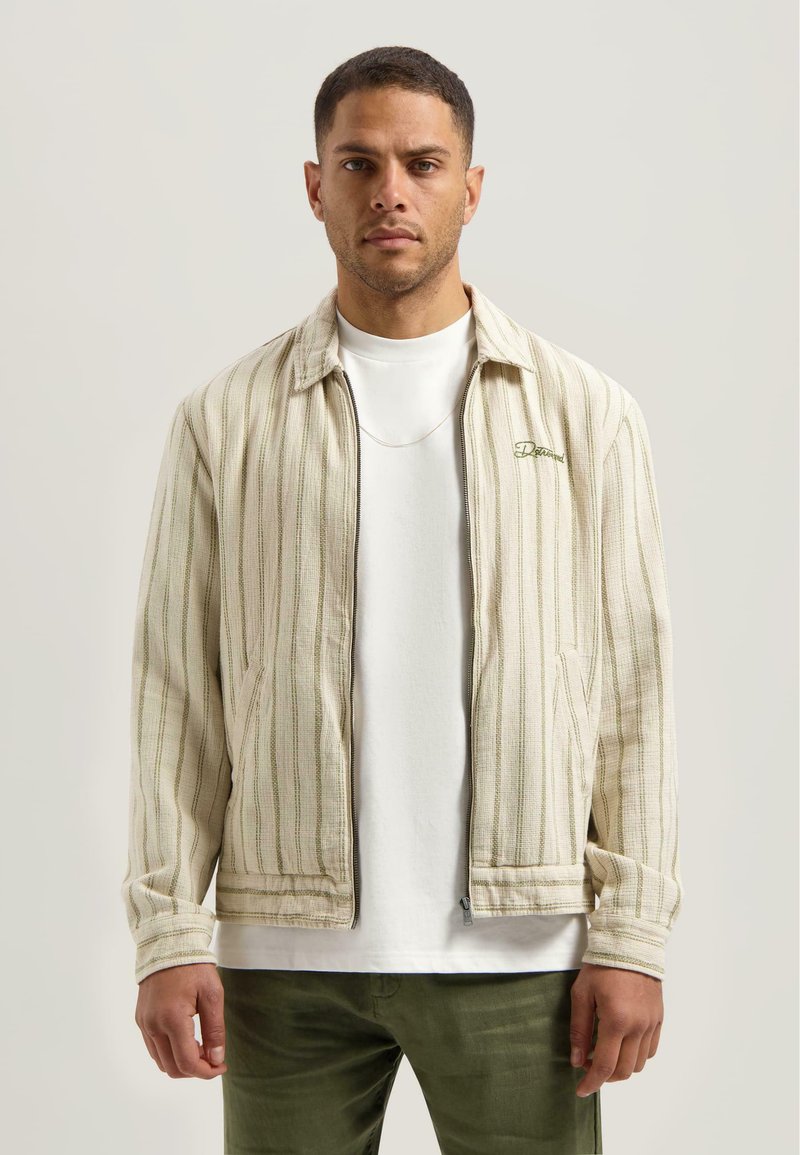 Chaqueta a rayas beige claro con detalles en verde, cierre de cremallera, bolsillos laterales y un cuello casual sobre una camisa blanca y pantalones verde oliva.