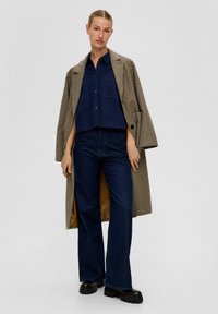 Gecuide lange jas over een marineblauw cropped shirt en donkerblauwe flared jeans. Zwarte laarzen met dikke zolen maken de outfit compleet.