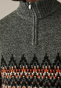 Grijze gebreide trui met een hoge kraag, voorzien van een zigzagpatroon in zwart, wit en oranje aan de onderrand. Ritssluitingdetail bij de hals.