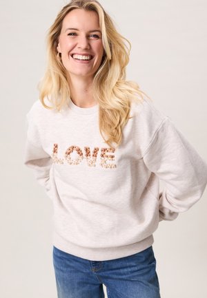 Lachende vrouw met lang blond haar, die een lichtbeige sweater draagt met het woord "LOVE" in parel- en paillettenletters en een blauwe spijkerbroek, met haar handen achter haar rug.