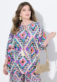 Mehrfarbige Bluse mit Aztekenmustern in Lila, Pink und Blau, mit ¾-Ärmeln, einem Schlüsselaugen-Ausschnitt und einem Quastenakzent. Goldene Handtasche mit Kettenriemen.