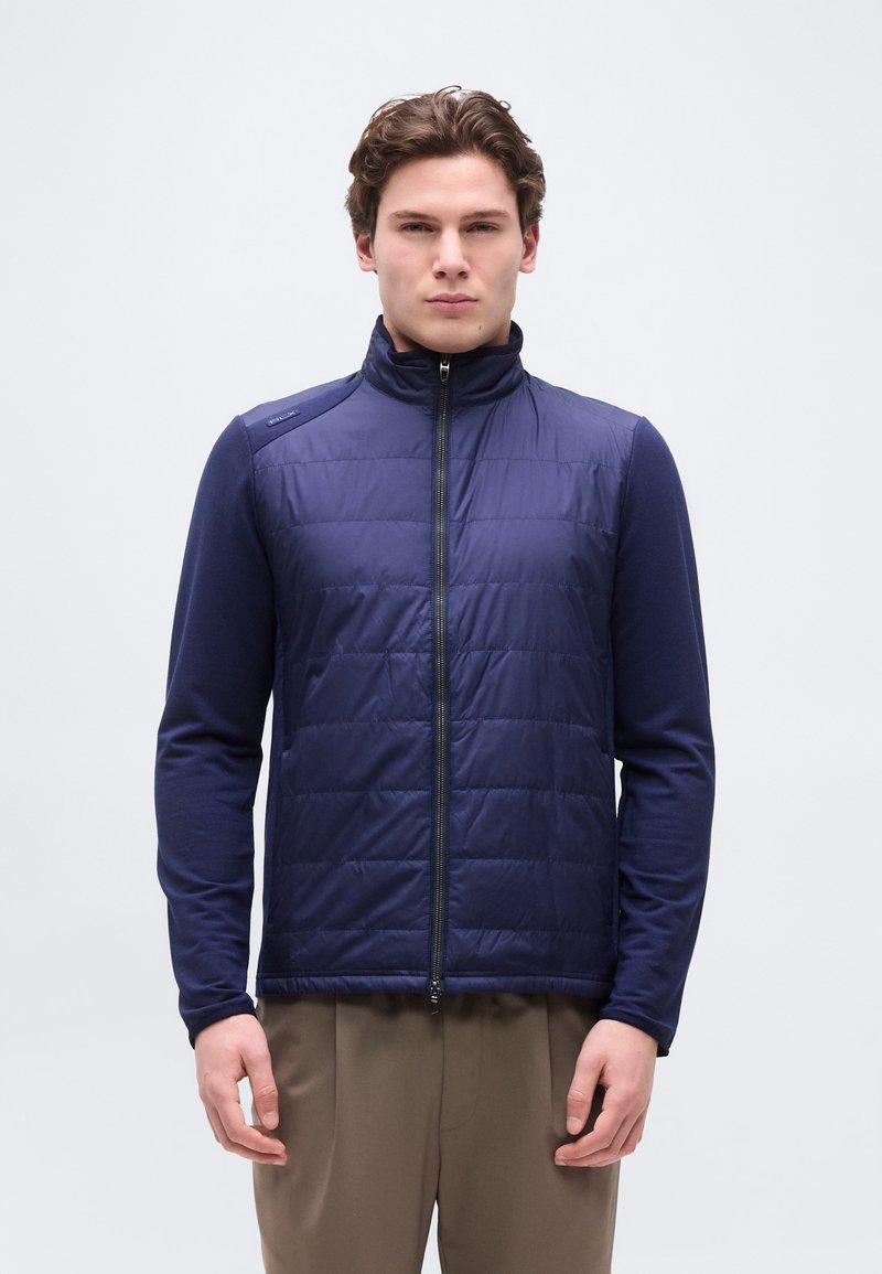 Νέος άνδρας που φοράει κοντό αμάνικο γιλέκο σε σ κούρο μπλε χρώμα πάνω από μακρυμάνικο πουκάμισο με μπεζ παντελόνι, στέκεται μπροστά σε ένα απλό φωτεινό φόντο.