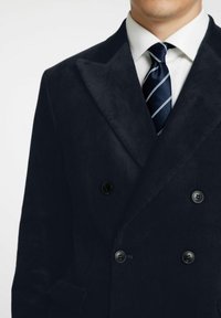 Marinblå kordflöjels dubbelknäppt jacka med breda slag, två svarta knappar och en vit skjorta med en marinblå och vit randig slips.