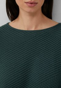 Donna che indossa un maglione lavorato a maglia verde scuro con un motivo a coste diagonali, mostrando il collo e la parte inferiore del viso con labbra naturali.