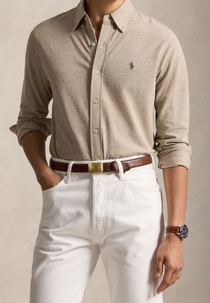 Homme portant une chemise beige à manches longues boutonnée, rentrée dans un pantalon blanc avec une ceinture en cuir marron et une montre-bracelet avec un bracelet marron.