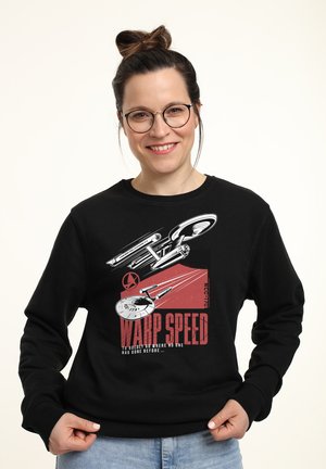 Uśmiechnięta kobieta w okularach w czarnej bluzie z kapturem z nadrukiem statku kosmicznego i czerwonym tekstem „WARP SPEED To boldly go where no one has gone before.”