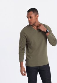 Olivegrünes Langarm-V-Ausschnitt-Shirt aus weichem Material. Model trägt dunkle Jeans. Uhr am linken Handgelenk, Hand berührt den Kragen.