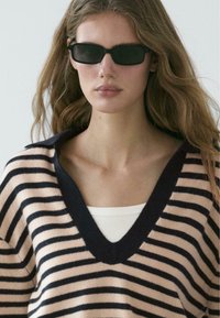 Maglione lavorato a maglia con scollo a V profondo, caratterizzato da strisce orizzontali nere e rosa. Il modello indossa occhiali da sole neri e una maglietta bianca sotto.