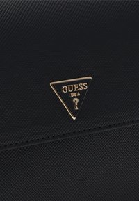 Svart texturerad handväska med en guldfärgad triangulär logotypplatta med texten "GUESS USA." Ytan visar ett subtilt mönster.