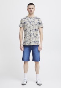 Beige kortermet t-skjorte med marineblå palme-design, kombinert med blå denimshorts. Modellen har på seg hvite sokker og grå joggesko med svarte detaljer.