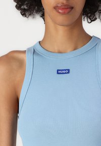Svetlo modrý rebrovaný top s okrúhlym výstrihom a modrou štítkou "HUGO" na prednej strane, vyrobený z mäkkého a elastického materiálu.