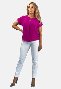 Fuksjowa bluzka z krótkim rękawem i okrągłym dekoltem, z detalem w postaci guzików, w zestawieniu z dopasowanymi białymi spodniami i wzorzystymi szpilkami.