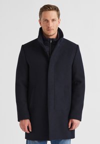 Mann, der einen dunkelblauen Wollmantel mit hohem Kragen über einer gepolsterten Jacke mit Reißverschluss und blauen Jeans trägt, vor einem schlichten weißen Hintergrund steht.