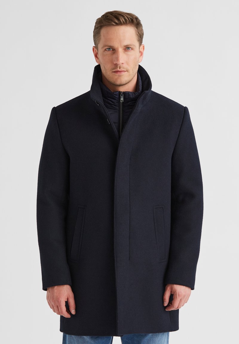 Mann, der einen dunkelblauen Wollmantel mit hohem Kragen über einer gepolsterten Jacke mit Reißverschluss und blauen Jeans trägt, vor einem schlichten weißen Hintergrund steht.