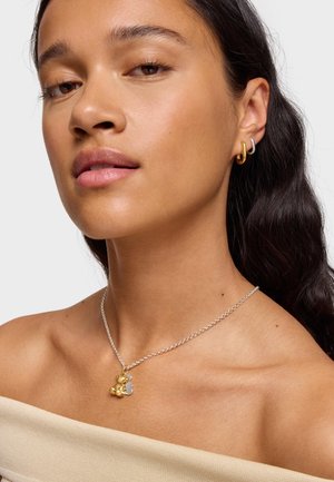 Die Halskette mit Anhänger aus Gold und Silber verfügt über runde und Blumen-Charms. Das Modell trägt eine Silberkette und goldene Creolen mit glatter Oberfläche.