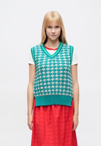 Bezrękawnik dziany w kolorze morskim i szarym, z teksturowanym wzorem diamentowym, dekoltem w serek i prążkowanym dołem. Noszony na białej koszulce.