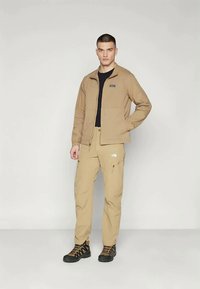 Beige letvægtsjakke og beige bukser, begge med en glat tekstur. Sort skjorte under. Sorte og orange vandresko med et robust design.