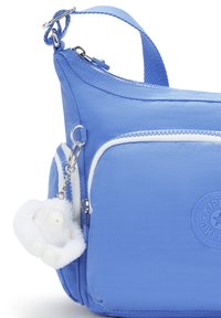 Blue nylon crossbody bag with white zipper accents and a circular logo. Features a soft, plush charm attached via a silver keyring.