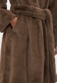 Hnedý župan z umelého kožušina s jemnou textúrou, širokými rukávmi a zodpovedajúcim opaskom. Obsahuje bočné vrecká pre väčšiu funkčnosť.