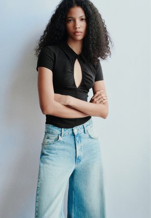Jeune femme aux cheveux bouclés portant un haut noir à manches courtes et un jean bleu clair, debout les bras croisés devant un mur clair uni.