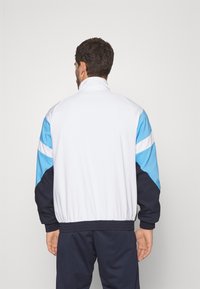 Atletiekjack met een witte romp, blauwe en marineblauwe mouwen, hoge kraag, elastische manchetten en tailleband. Achteraanzicht toont het kleurblokontwerp.