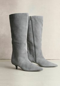 Pair of gray suede knee-high boots with pointed toes, small kitten heels, and visible side zippers, standing on a neutral surface.