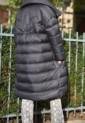 Persona vistiendo un abrigo negro largo acolchado con capucha sobre pantalones estampados, de pie cerca de una cerca de metal con arbustos verdes detrás.