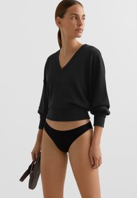 Čierna mikina s výstrihom do V, dlhými rukávmi a rebrovanými manžetami, kombinovaná s čiernymi rebrovanými bikinovými spodkami. Modelka drží tmavo hnedú tašku.