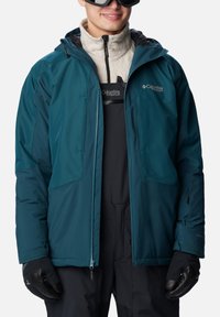 Türelblau Winterjacke mit schwarzen Akzenten, ausgestattet mit zwei Brusttaschen, einem Reißverschluss vorne und einem hohen Kragen. Getragen über einer leichten isolierenden Schicht.
