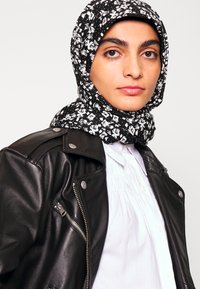 Jaqueta de motoqueiro de couro negro com fechos prateados, sobre uma camisa branca com colarinho. O lenço negro apresenta um padrão floral branco.