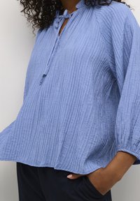 Blouse bleu clair à texture froissée avec un col rond et une fermeture à nouer. Manches trois quarts avec poignets froncés.