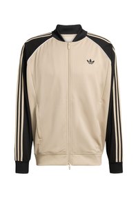 Beež sportjakk võrkmustriga, mustade detailide ja kolme musta triibuga varrukaosal. Varustatud luku ja musta kraega.