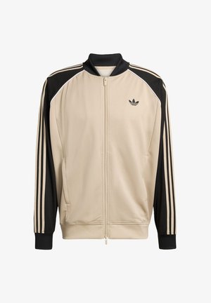 Bež sportska jakna s mrežastom teksturom, crnim akcentima i tri crne pruge na rukavima. Ima zatvarač na patent i crni ovratnik.