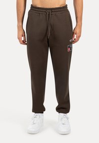Pantalones de chándal marrones de algodón con cintura elástica y cordón, con una etiqueta lateral y dobladillos ajustados. Combinados con zapatillas blancas.