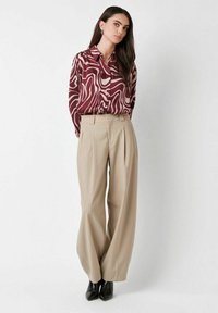 Woman wearing a maroon and beige abstract patterned button-up shirt tucked into wide-leg beige trousers and black pointed ankle boots.