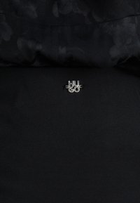 Silberner "HU" und "60" Logo-Anstecker auf glattem schwarzen Stoff, zentriert, umgeben von einem strukturierten schwarzen Kleidungsstück mit floralen Mustern.