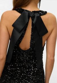 Juodas blizgučiais puoštas suknelė su kaklo juostos iškirpte, kurią puošia didelė juoda juosta. V formos nugaros iškirptė atidengia odą. Blizgūs blizgučiai sukuria tekstūruotą vaizdą.