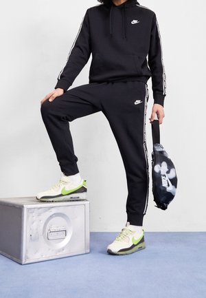 Osoba w czarnym dresie Nike z białymi paskami stoi jedną stopą na metalowej skrzynce, trzymając czarno-białą torbę Nike, ubrana w biało-zielone sneakersy.