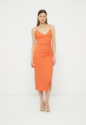 Kvinde stående iført en ærmeløs orange midi-kjole med en sideslids og beige ankelstrophæle mod en ensfarvet hvid baggrund.