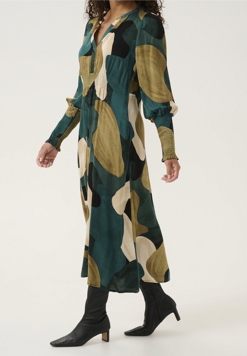Femme portant une longue robe à imprimé abstrait avec des manches froncées et des bottines noires à talons moyens, debout devant un fond uni.