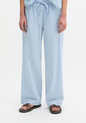 Persoon draagt lichtblauwe wijde broek met trekkoord in de taille en donkerbruine platte sandalen op een witte achtergrond.