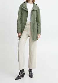 Parka verde leggera con cappuccio regolabile, zip frontale e bottoni; abbinata a pantaloni ampi color crema e stivali neri con tacchi e dettagli bianchi.