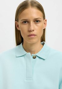 Hellblaues Poloshirt mit einem geknöpften Kragen, hergestellt aus weichem Stoff. Es zeichnet sich durch eine dezente Textur und ein einzelnes Messingknopf-Detail aus.