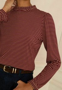 Dug prašnjav i kremasti prugasti dugih rukava s ruffles-om na lažnom ovratniku i napuhanim rukavima, uparen s smešnim remenom i tamnim trapericama.