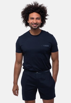 Mann mit lockigem Haar, lächelnd, trägt ein dunkelblaues T-Shirt und passende Shorts, steht mit einer Hand in der Tasche vor weißem Hintergrund.