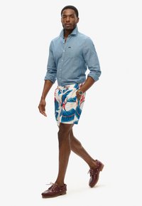 Camisa de chambray azul claro con mangas arremangadas, pantalones cortos de baño blancos con estampado floral que presentan hojas azules y rojas, y zapatos de barco marrón oscuro.